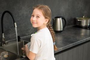 cute little girl with a pigtail washes dishes in the kitchen at the sink and smiles photo
