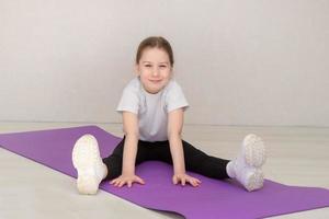 una niña linda con uniforme deportivo se sienta en una alfombra de gimnasia con los brazos extendidos hacia adelante y sonríe foto