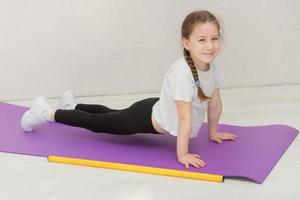 cute little girl stands in a plank on a mat, a gymnastic stick lies nearby, the child smiles photo