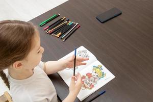 niña pequeña con una coleta sentada en la mesa elige lápices para colorear según el modelo foto