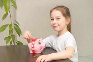 financial literacy concept cute little girl sitting at the table throws a coin into the piggy bank and smiles photo