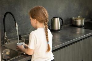 linda niñita con coleta lava platos en la cocina en el fregadero foto