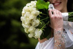 The bride in a white wedding dress is holding a bouquet of white flowers - peonies, roses. Wedding. Bride and groom. Delicate welcome bouquet. Beautiful decoration of weddings with leaves photo