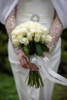 The bride in a white wedding dress is holding a bouquet of white flowers - peonies, roses. Wedding. Bride and groom. Delicate welcome bouquet. Beautiful decoration of weddings with leaves photo