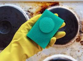 hand in household glove holding sponge with cleaning liquid over dirty stove photo