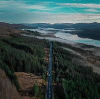 A straight road makes its way through a stunning Scottish highland area. The photo is taken from a drone's perspective, giving a bird's eye view of the rolling hills and picturesque landscape.