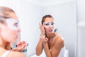 Woman applying mask moisturizing skin mask on face looking in bathroom mirror. Girl taking care of her complexion layering moisturizer. Skincare spa treatment. photo