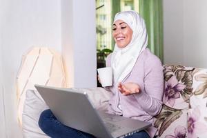 Muslim woman working with computer. Arab Young business woman sitting at her desk at home, working on a laptop computer and drinking coffee or tea. Muslim woman working at a home and using computer. photo