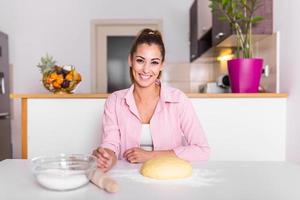 hermosa joven mirando a la cámara y sonriendo mientras hornea en la cocina en casa. sonriente joven ama de casa haciendo masa foto