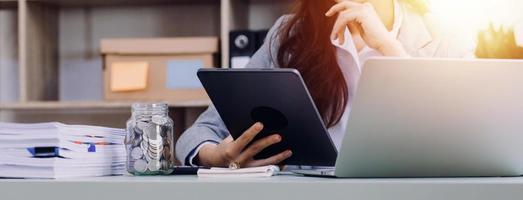 Young business woman sitting in office at table and using smartphone. On desk is laptop and tablet computer, on screen charts and graphs. Woman analyzing data. Student learning online. photo