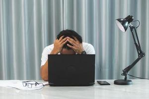 Stressful office job finally turns young man into a burnout. Stressed, tired, exhausted male worker overwhelmed with problems sitting at working desk with laptop computer and holding hand on his face photo