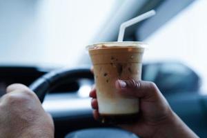la conductora asiática sostiene una taza de café con hielo y pan de sándwich para comer y beber en el automóvil, es peligrosa y corre el riesgo de sufrir un accidente. foto