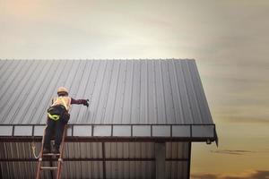 trabajador de techado con uniforme protector y guantes, herramientas para techos, instalación de techos nuevos en construcción, taladro eléctrico usado en techos nuevos con láminas de metal. foto