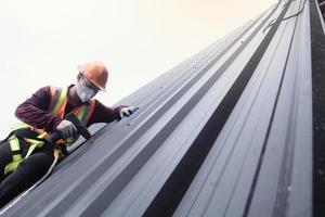 trabajador de techado con uniforme protector y guantes, herramientas para techos, instalación de techos nuevos en construcción, taladro eléctrico usado en techos nuevos con láminas de metal. foto