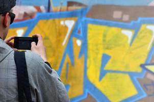 un joven grafitero fotografía su cuadro completo en la pared. el chico usa tecnología moderna para capturar un colorido dibujo de graffiti abstracto. centrarse en el dispositivo de fotografía foto