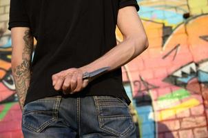 vista trasera de un joven caucásico con un cuchillo en la mano contra la pared de ladrillo del ghetto con pinturas de graffiti. concepto de fuerzas criminales y cargo de agresión foto