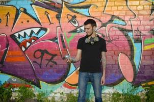 Young caucasian graffiti artist in black t-shirt with silver aerosol spray can near colorful graffiti in pink tones on brick wall. Street art and contemporary painting process photo