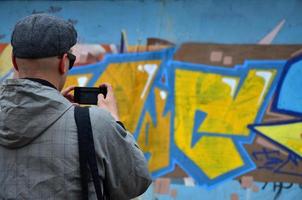 un joven grafitero fotografía su cuadro completo en la pared. el chico usa tecnología moderna para capturar un colorido dibujo de graffiti abstracto. centrarse en el dispositivo de fotografía foto