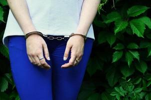 fragmento del cuerpo de una joven criminal con las manos esposadas contra un fondo verde de hojas de hiedra floreciente. el concepto de detener a un delincuente de una mujer criminal en un entorno rural foto