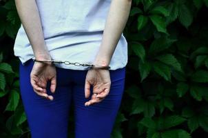 Fragment of a young criminal girl's body with hands in handcuffs against a green blossoming ivy leaves background. The concept of detaining an offender of a female criminal in a rural environment photo