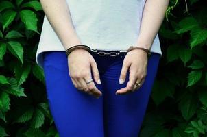 fragmento del cuerpo de una joven criminal con las manos esposadas contra un fondo verde de hojas de hiedra floreciente. el concepto de detener a un delincuente de una mujer criminal en un entorno rural foto