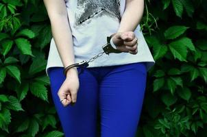 Fragment of a young criminal girl's body with hands in handcuffs against a green blossoming ivy leaves background. The concept of detaining an offender of a female criminal in a rural environment photo