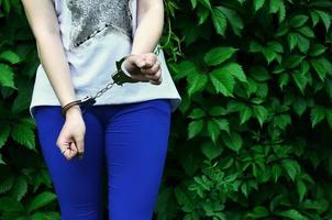 Fragment of a young criminal girl's body with hands in handcuffs against a green blossoming ivy leaves background. The concept of detaining an offender of a female criminal in a rural environment photo