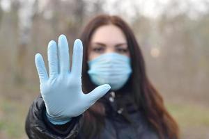 retrato de una joven morena con máscara protectora azul y guantes de goma muestra un gesto de parada al aire libre en madera de primavera foto