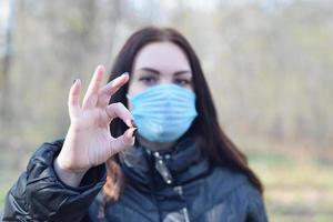 Portrait of young brunette woman in blue protective mask shows okay gesture outdoors in spring wood photo