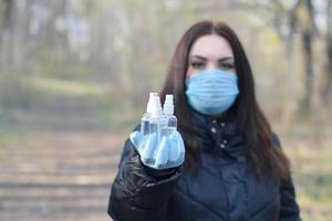 mujer joven con máscara protectora muestra botellas de spray desinfectante al aire libre en madera de primavera foto