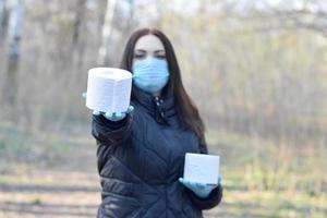 Covidiot concept. Young woman in protective mask gives roll of toilet paper outdoors in spring wood photo