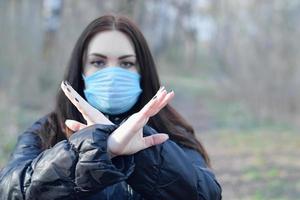 Portrait of young brunette woman in blue protective mask shows stop gesture outdoors in spring wood photo