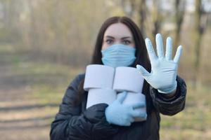 concepto covidiota. mujer joven con máscara protectora sostiene muchos rollos de papel higiénico y muestra un gesto de parada al aire libre en madera de primavera foto