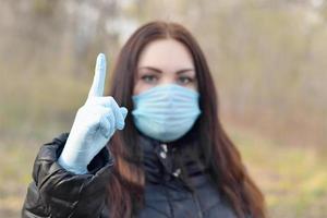 Portrait of young brunette woman in blue protective mask and rubber gloves shows attention gesture outdoors in spring wood photo