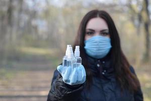 mujer joven con máscara protectora muestra botellas de spray desinfectante al aire libre en madera de primavera foto