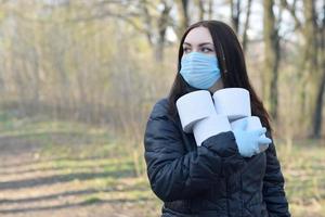 Covidiot concept. Young woman in protective mask holds many rolls of toilet paper outdoors in spring wood photo