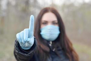 Portrait of young brunette woman in blue protective mask and rubber gloves shows attention gesture outdoors in spring wood photo