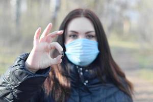 Portrait of young brunette woman in blue protective mask shows okay gesture outdoors in spring wood photo