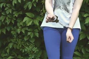 Fragment of a young criminal girl's body with hands in handcuffs against a green blossoming ivy leaves background. The concept of detaining an offender of a female criminal in a rural environment photo