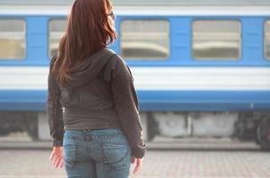 una joven pelirroja está parada en la plataforma ferroviaria y observa el tren que sale. la mujer llegaba tarde a su tren. vista trasera foto