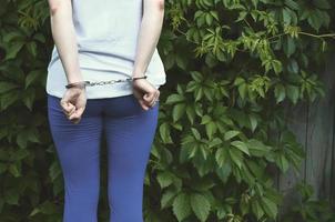 Fragment of a young criminal girl's body with hands in handcuffs against a green blossoming ivy leaves background. The concept of detaining an offender of a female criminal in a rural environment photo