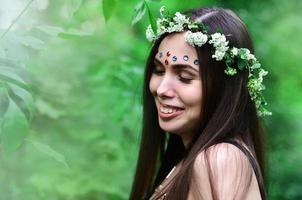 retrato de una joven emocional con una corona floral en la cabeza y adornos brillantes en la frente. linda morena posando en un hermoso bosque floreciente durante el día en un buen día foto