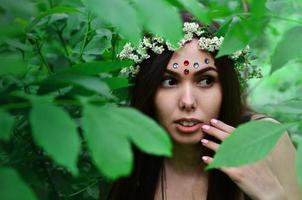 una foto del bosque de una hermosa joven morena de apariencia europea con ojos marrones oscuros y labios grandes. en la cabeza de la niña lleva una corona de flores, en la frente adornos brillantes