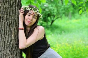 retrato de una joven emocional con una corona floral en la cabeza y adornos brillantes en la frente. linda morena posando en un hermoso bosque floreciente durante el día en un buen día foto