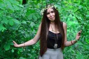 una foto del bosque de una hermosa joven morena de apariencia europea con ojos marrones oscuros y labios grandes. en la cabeza de la niña lleva una corona de flores, en la frente adornos brillantes