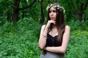 A forest picture of a beautiful young brunette of European appearance with dark brown eyes and large lips. On the girl's head is wearing a floral wreath, on her forehead shiny decorations photo