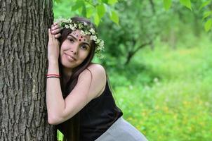 Portrait of an emotional young girl with a floral wreath on her head and shiny ornaments on her forehead. Cute brunette posing in a burgeoning beautiful forest in the daytime on a fine day photo