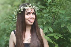 Portrait of an emotional young girl with a floral wreath on her head and shiny ornaments on her forehead. Cute brunette posing in a burgeoning beautiful forest in the daytime on a fine day photo