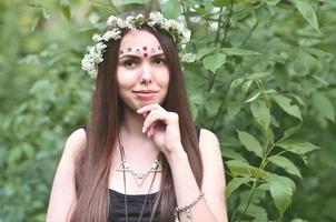 retrato de una joven emocional con una corona floral en la cabeza y adornos brillantes en la frente. linda morena posando en un hermoso bosque floreciente durante el día en un buen día foto