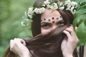 retrato de una joven emocional con una corona floral en la cabeza y adornos brillantes en la frente. linda morena posando en un hermoso bosque floreciente durante el día en un buen día foto